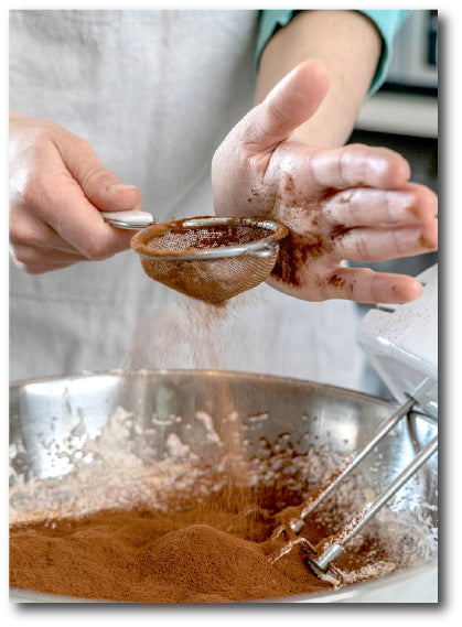 Dementia-friendly reminiscence image of a chef preparing a chocolate cake mix by sieving cocoa powder into a bowl of other ingredients ready for mixing 