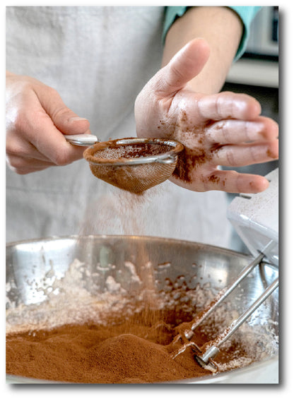 Dementia-friendly reminiscence image of a chef preparing a chocolate cake mix by sieving cocoa powder into a bowl of other ingredients ready for mixing 
